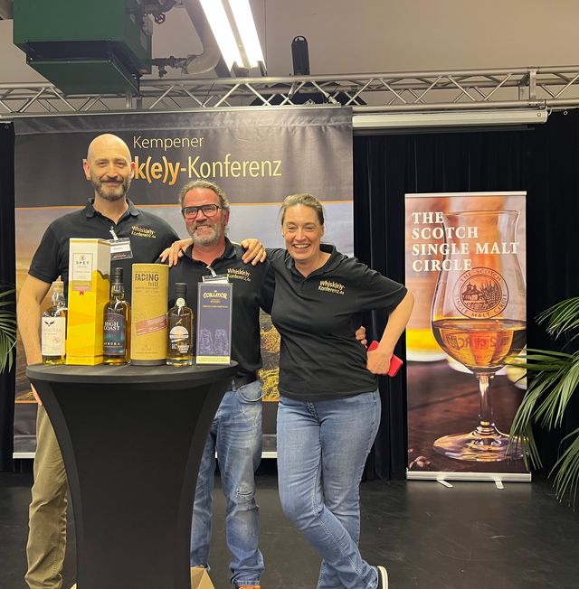 Drei Personen mit Whiskyproben vor Banner.